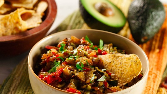 Chunky grilled vegetable guacamole in bowl with tortillas and avocado in background