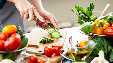 Person slicing a cucumber on a wooden cutting board surrounded by fresh vegetables.